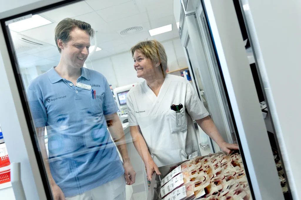 Två personer, en i blå tröja med texten ”Region Holland” och en i vit vårduniform, står och pratar. Ett skåp med blodpåsar syns i förgrunden.