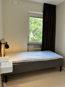 Sovrum med säng, lampa, sängbord och fönster.